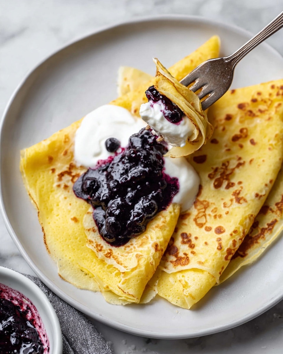 A white plate holds three thin, folded yellow crepes with light brown spots, arranged overlapping each other. On top, there is a dollop of white creamy yogurt, partially covered by a thick, dark purple blueberry sauce. A fork is lifting a small rolled piece of crepe that has blueberry sauce inside. The scene sits on a white marbled surface with a part of a white bowl containing more blueberry sauce visible at the bottom left corner. photo taken with an iphone --ar 4:5 --v 7