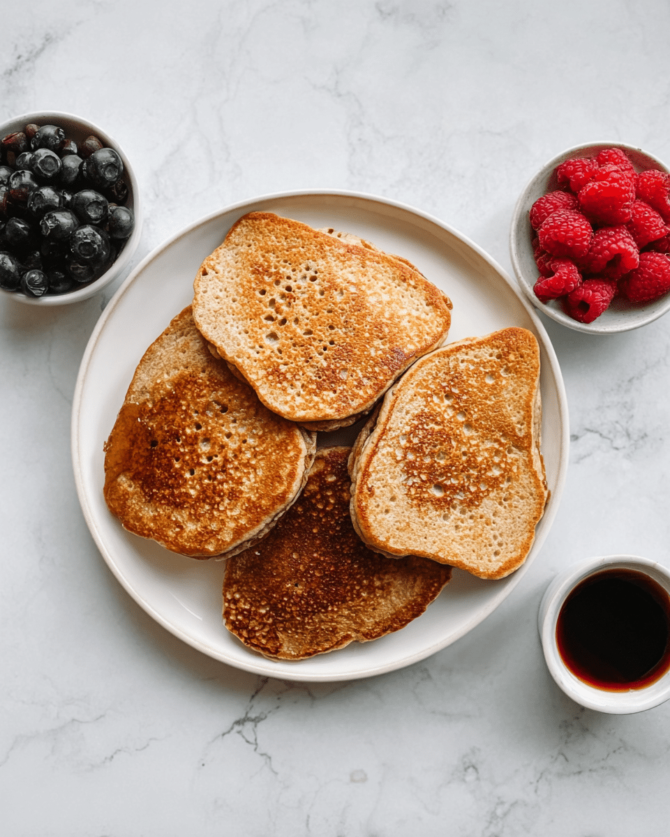 A white plate holds four thick, light brown pancakes, each with a slightly uneven shape and a golden-brown surface showing round patterns and small bubbles. The pancakes are stacked loosely, overlapping each other. To the right of the plate, there is a small white bowl filled with fresh red raspberries, and above it, a similar bowl with dark blue blueberries. Below the plate, a small white cup contains a dark syrup. All items are placed on a white marbled texture surface. Photo taken with an iphone --ar 4:5 --v 7