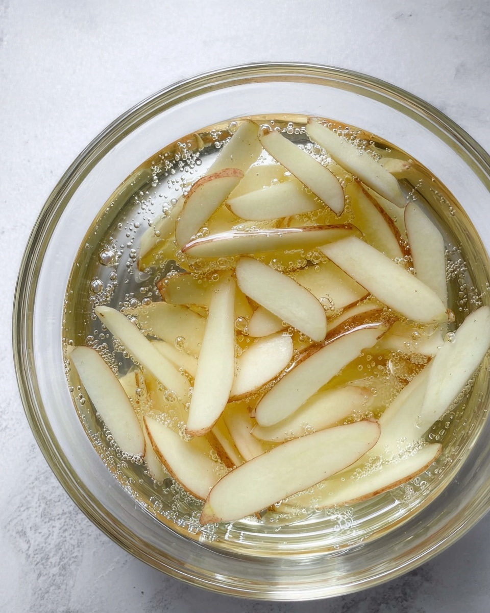 A clear glass bowl is filled with long, thin white potato slices with some brown skin still on them, all soaking in clear water with tiny bubbles. The bowl sits on a white marbled surface, and the potato pieces float in different directions inside the water. photo taken with an iphone --ar 4:5 --v 7