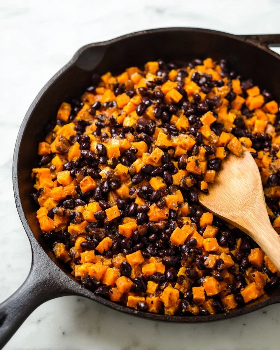 A close-up image of a black cast iron pan filled with a mix of small, bright orange cubes and shiny black beans evenly spread inside. The orange cubes have a soft texture with light browning on their edges, while the black beans provide a smooth, rounded contrast throughout. A light brown wooden spoon is resting on the right side of the pan, partially mixing the contents. The pan sits on a white marbled surface, giving a clean and bright backdrop to the warm colors of the food photo taken with an iphone --ar 4:5 --v 7