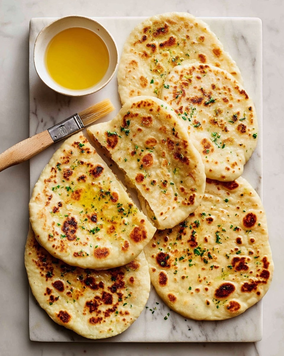 The image shows five flat, round pieces of naan bread arranged on a white marbled surface. Each naan has a golden-brown toasted texture with some darker charred spots and light bubbling on the surface, sprinkled with small bits of green herbs. One piece is folded in half, showing its soft inside. On the left side, there is a small white bowl filled with melted butter or oil, next to a wooden-handled brush resting on the marble. The colors are light yellows and browns, and the surface is clean and bright. photo taken with an iphone --ar 4:5 --v 7
