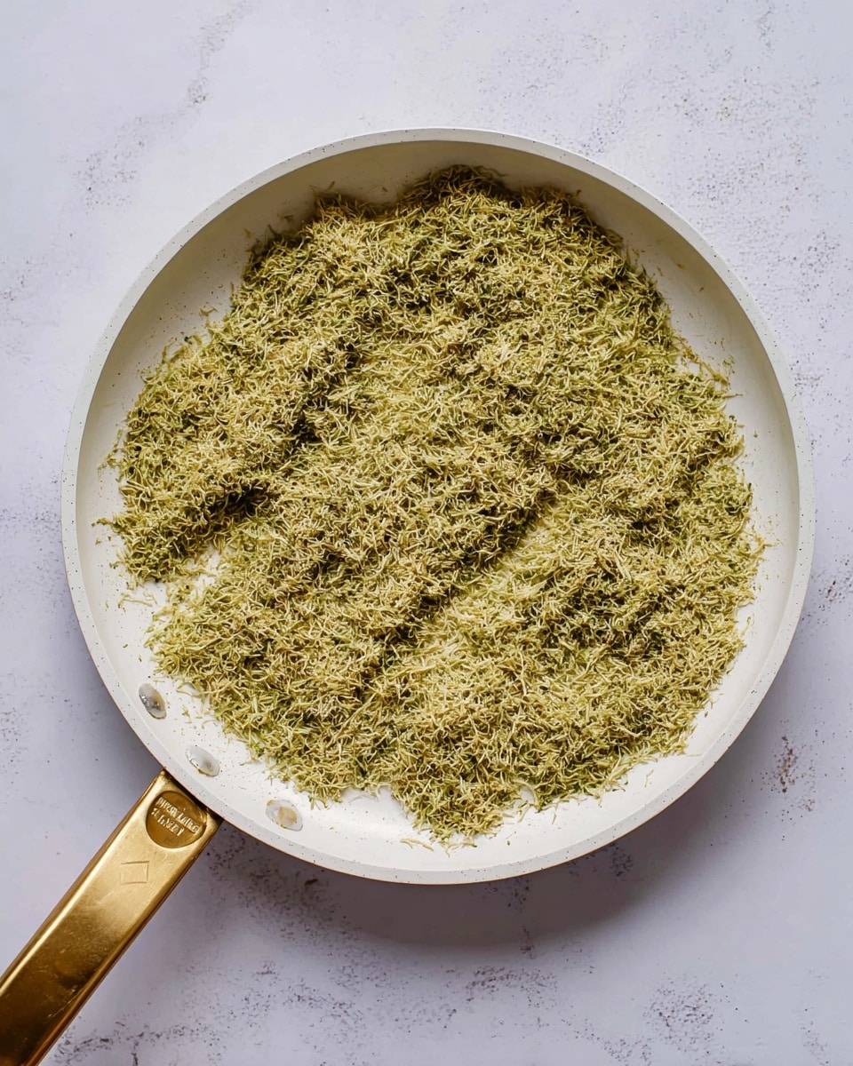 A white frying pan with a golden handle holds a single thick layer of greenish-yellow fine shredded herb mixture spread evenly across the bottom. The mixture has a fibrous texture with thin strands visible all over. The pan sits on a white marbled surface, with some stray strands scattered lightly around the edges of the pan. photo taken with an iphone --ar 4:5 --v 7