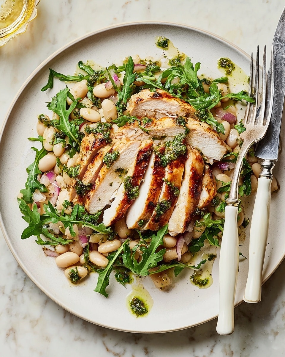 A white plate holds a dish with three layers: the bottom layer is a bed of white beans mixed with small pieces of red onion and scattered green arugula leaves, creating a fresh look. On top are several slices of grilled chicken with a golden-brown, slightly charred crust and a white tender inside, arranged in a slight overlap across the center. The dish is drizzled with a green herb sauce adding bright spots of color, and a fork and knife with white handles rest on the right edge of the plate. The plate sits on a white marbled textured surface. Photo taken with an iphone --ar 4:5 --v 7