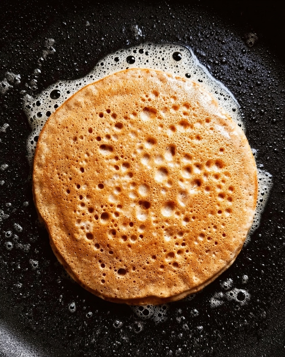 A single round pancake with a light brown color is cooking in a black pan. The pancake has many small holes and bubbles on the surface, with a slightly uneven texture showing specks of darker brown throughout. Around the pancake, there are small foamy bubbles of oil or butter on the pan, giving a shiny look. The pancake edges are soft and slightly raised from the pan. photo taken with an iphone --ar 4:5 --v 7