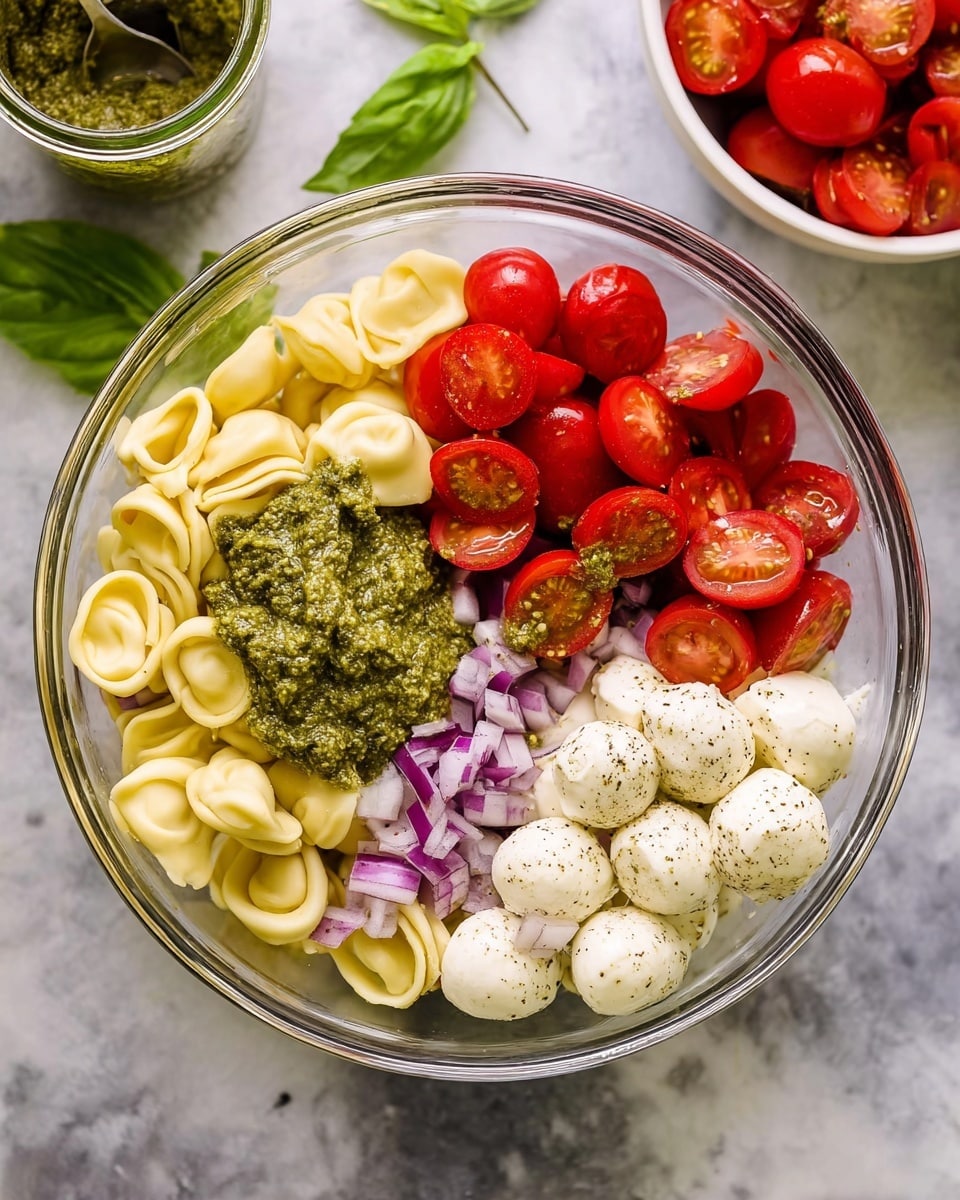 A clear glass bowl sits on a white marbled surface filled with ingredients for a fresh pasta salad. Inside the large bowl, on the left side, there are smooth, pale yellow tortellini pasta pieces forming the base layer. On top of the pasta, on the upper left, there is a thick, light green pesto sauce with a slightly coarse texture. The top right section of the bowl is packed with shiny, halved red cherry tomatoes. The bottom right part holds round white mozzarella balls lightly speckled with black pepper and herbs. In the bottom left corner, there are finely chopped small pieces of purple-red onion layered next to the mozzarella. A fresh green basil leaf rests near the onion. In the blurred background, a small white bowl filled with halved cherry tomatoes sits next to an open jar of pesto with a spoon inside, and some fresh green basil leaves are partially visible. photo taken with an iphone --ar 4:5 --v 7