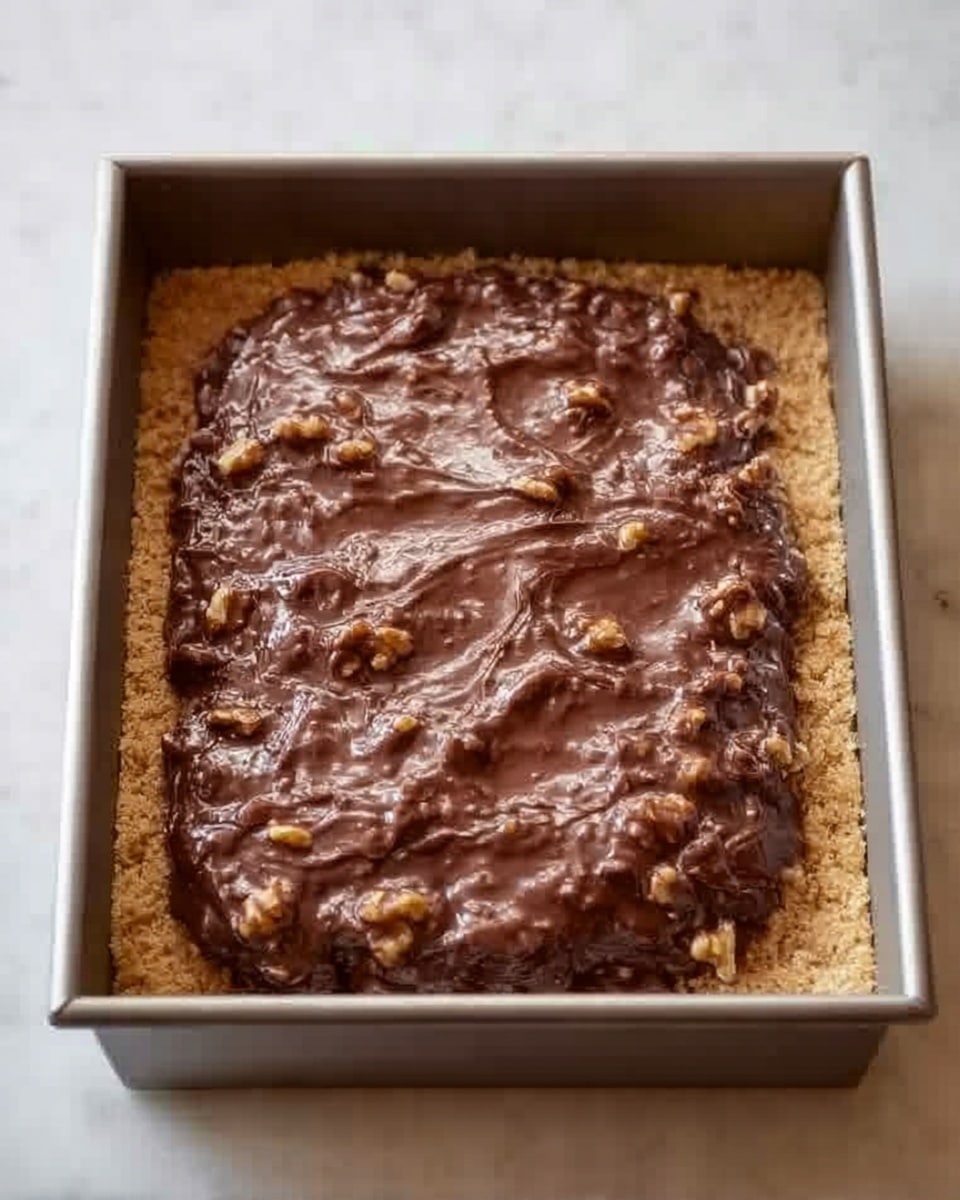 A square metal baking pan holds a layered dessert set on a white marbled surface. The bottom layer is a light golden crumb crust, smooth and even, filling the pan. On top is a thick, uneven layer of rich dark brown chocolate fudge with walnut pieces mixed in, creating a bumpy texture across the surface. The edges of the dessert show where the fudge layer spreads slightly over the crust. Photo taken with an iphone --ar 4:5 --v 7