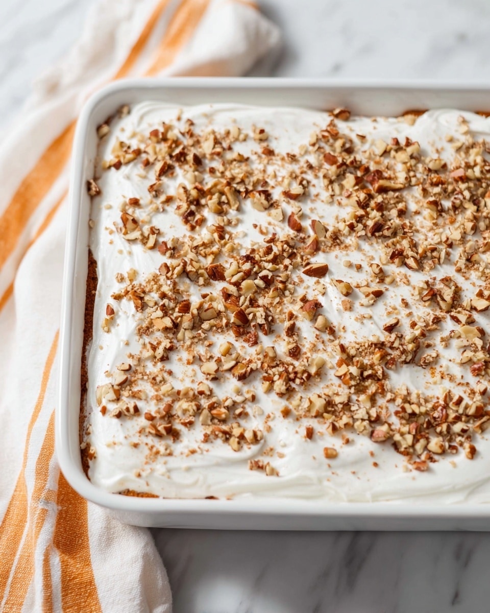 A rectangular white dish filled with a dessert that has two visible layers; the bottom layer is not clearly seen but is covered by a thick, smooth white layer of frosting on top. The white frosting layer is spread evenly with a light texture and sprinkled generously with small pieces of chopped brown nuts across the whole surface. The dish is set on a white marbled surface with a white towel showing orange stripes partially visible on the left side. Photo taken with an iphone --ar 4:5 --v 7