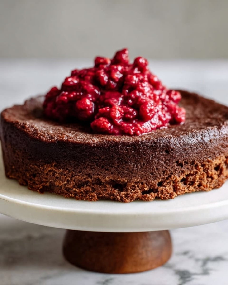 The image shows a single-layer round chocolate cake with a dark brown, slightly cracked and textured surface. On top of the cake, there is a dollop of bright red berry compote, which looks chunky and glossy with visible whole berries. The cake sits on a white plate with a wooden stand underneath, all placed on a white marbled texture surface. Photo taken with an iphone --ar 4:5 --v 7