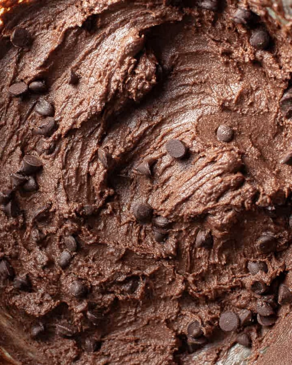 A close-up view of thick, dark brown chocolate dough mixed with small pieces of chocolate or tiny round chocolate bits scattered throughout. The texture looks soft and dense with a few shiny spots where the light hits the smooth, thick dough. The dough fills the frame and looks rich and moist, all set against a white marbled surface. photo taken with an iphone --ar 4:5 --v 7