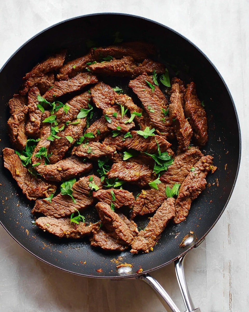 A black pan filled with cooked, thin slices of brown meat scattered evenly across the bottom, each piece showing a slightly rough texture and grill marks on some slices. Small green herb leaves are sprinkled on top, adding a fresh contrast to the rich brown meat. The pan’s handle extends out from the lower right, resting on a white marbled surface that serves as the background. photo taken with an iphone --ar 4:5 --v 7