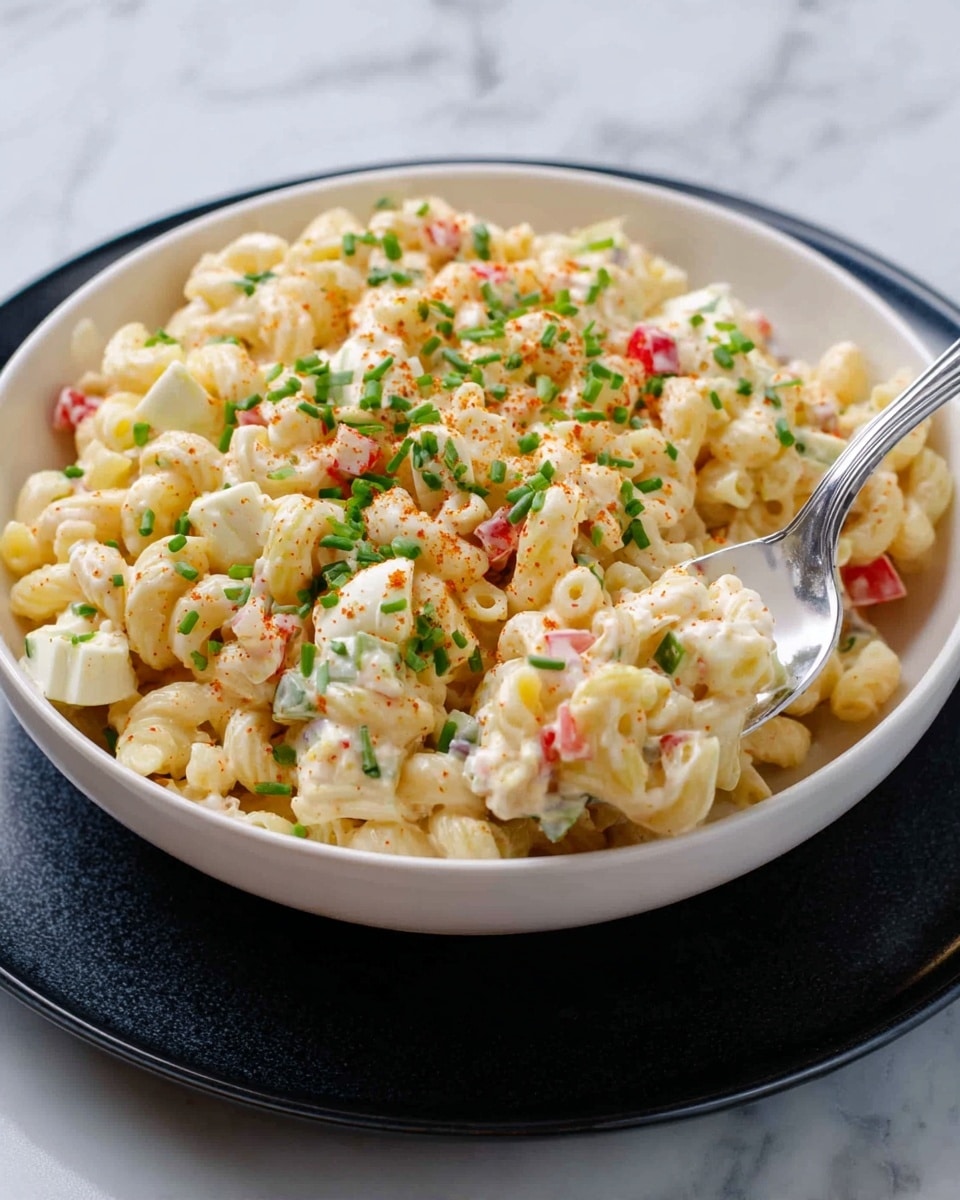 A white bowl filled with creamy macaroni salad, showing small elbow pasta pieces mixed with chopped hard-boiled eggs, diced red bell peppers, and green celery bits. The dish is topped with chopped fresh chives and a light sprinkle of paprika. A silver spoon rests inside the bowl on the right side, partially scooping the salad. The bowl sits on a black round plate, placed against a white marbled surface. photo taken with an iphone --ar 4:5 --v 7