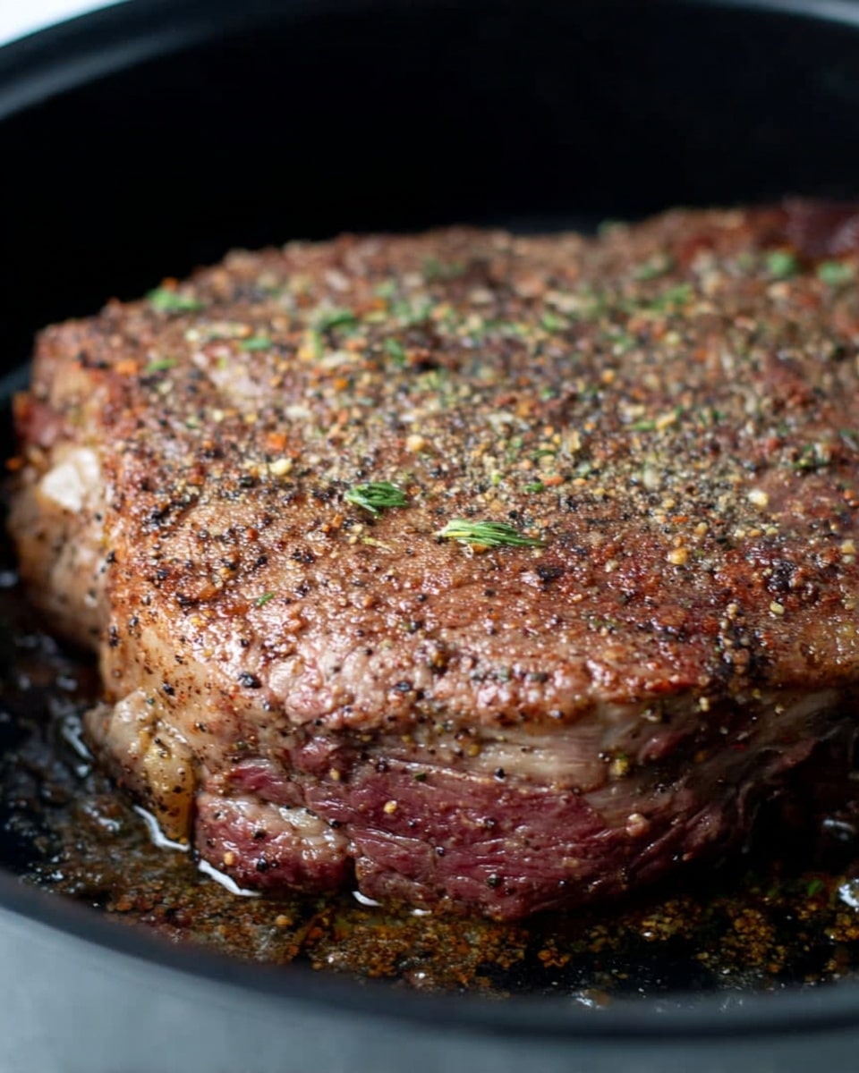 A close-up view of a thick, cooked steak in a black pan, showing a seasoned brown crust on top with visible coarse spices like black pepper and herbs spread evenly. The steak has a slightly pink edge where the meat meets the fat, and the texture is rough and juicy looking. The pan holds some oil around the edges of the steak, giving it a shiny appearance. The surface beneath the pan is a white marbled texture photo taken with an iphone --ar 4:5 --v 7
