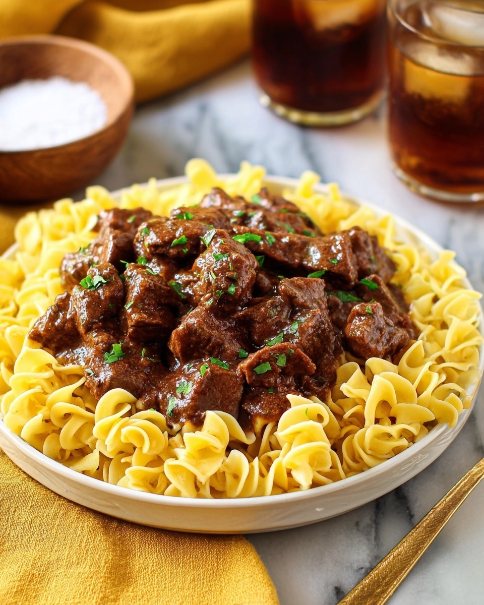 A white plate with yellow egg noodles forming the bottom layer, the noodles have a smooth, curled texture and are loosely piled, on top is a thick layer of dark brown beef stew pieces covered in a glossy rich sauce, the beef chunks look tender and are sprinkled with small bits of chopped green parsley, the plate sits on a white marbled surface with a yellow cloth and fork placed to the right side, a small wooden bowl with salt and iced tea glasses slightly blurred are in the background, photo taken with an iphone --ar 4:5 --v 7