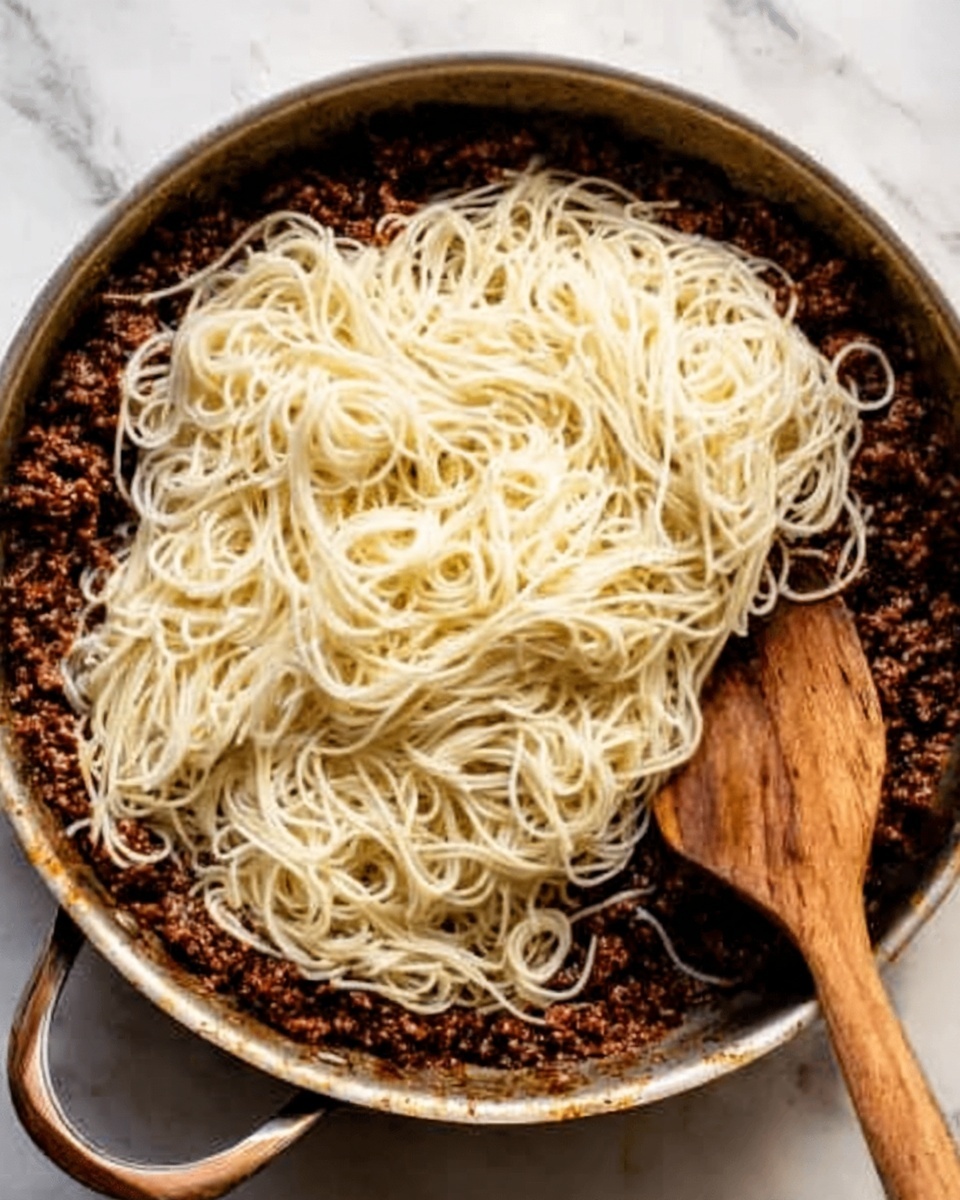 A pan filled with two layers: the bottom layer is dark brown cooked meat sauce with a textured, crumbly look, covering the whole pan base, and the top layer is a heap of smooth, soft, pale yellow noodles spread evenly over the meat. A wooden spatula is on the right side, partly submerged in the noodles and sauce. The pan sits on a white marbled surface. Photo taken with an iphone --ar 4:5 --v 7