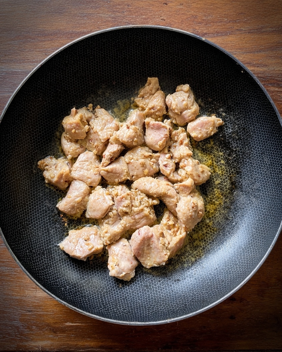 The image shows a black frying pan with a hexagon pattern on the inside, placed on a wooden surface. Inside the pan, there are multiple pieces of small light pinkish beige chicken cooking, with a slightly browned texture on some parts and a thin layer of oil around them. The chicken pieces look soft and juicy, and they cover most of the pan's middle area. The lighting highlights the different shades of the chicken and oil. Photo taken with an iphone --ar 4:5 --v 7