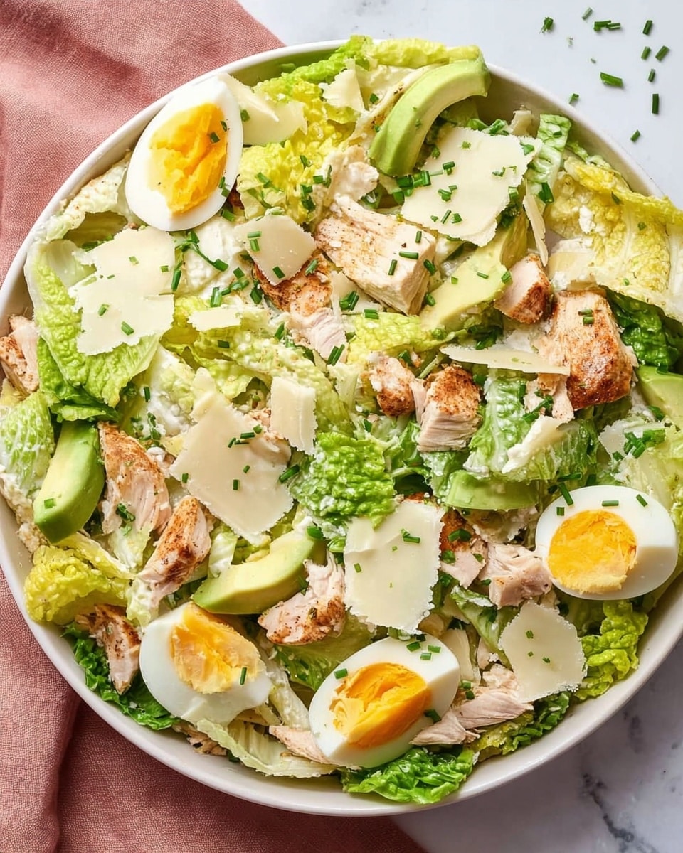 A white bowl filled with a fresh salad placed on a white marbled surface with a muted pink cloth nearby. The salad has several layers including bright green romaine lettuce leaves as the base, topped with sliced boiled eggs showing firm yellow yolks and white edges, chunks of cooked chicken with a light brown grilled texture, and large pale green avocado wedges. Scattered on top are thin, uneven shavings of a pale yellow cheese and finely chopped green chives sprinkled all over. The salad has a light dressing giving a slight sheen to the ingredients. photo taken with an iphone --ar 4:5 --v 7