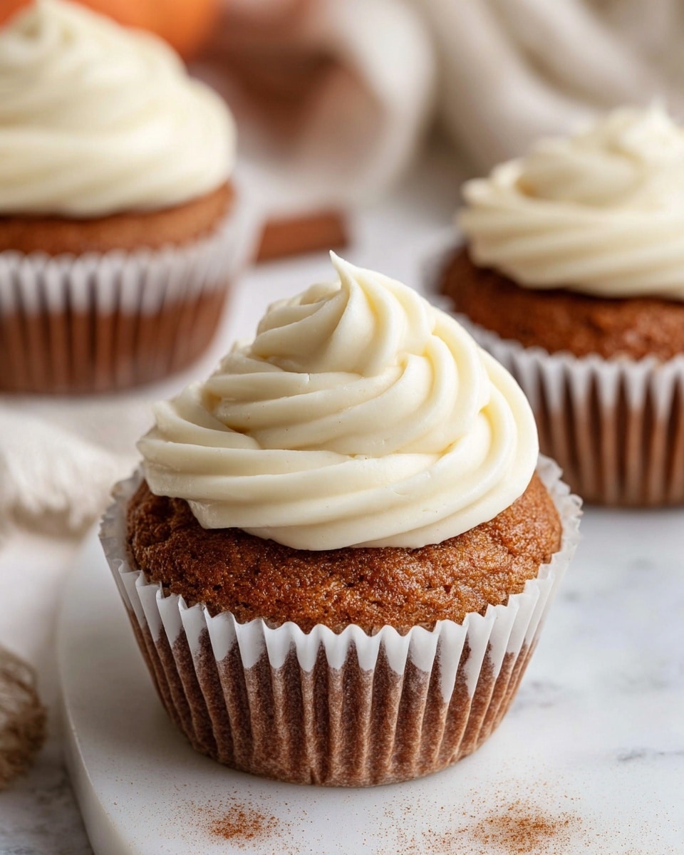 The image shows a close-up of a single cupcake with a brown, textured base and a thick swirl of smooth, white frosting on top. The cupcake sits in a white paper liner, slightly opened at the edges, placed on a white marble surface with some cinnamon powder sprinkled around. In the background, two more cupcakes are partially visible, each topped with the same creamy white swirl of frosting. The overall feel is soft and inviting, with warm brown and creamy white colors standing out. Photo taken with an iphone --ar 4:5 --v 7