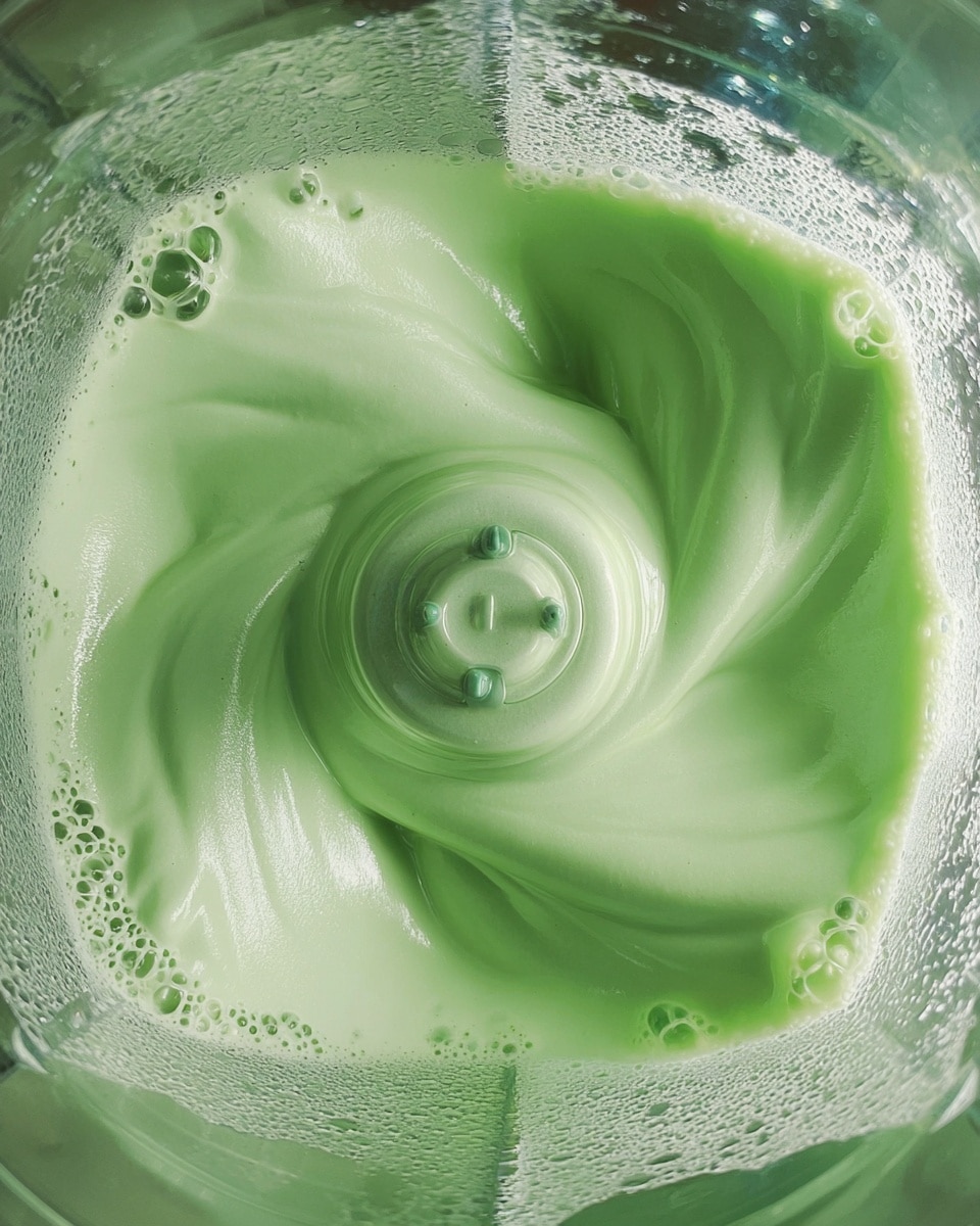 A close-up view inside a blender shows a thick, light green creamy mixture swirling around the blades in the center. The mixture has a smooth, foamy texture with bubbles forming along the edges, creating a dynamic motion effect. The light reflects softly on the smooth surface of the mixture as it spins in the clear blender container. photo taken with an iphone --ar 4:5 --v 7