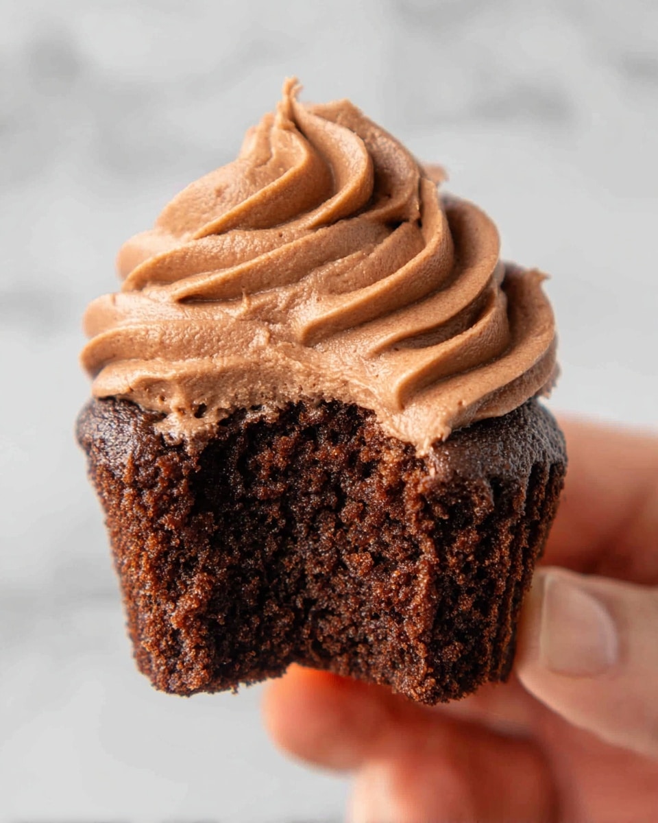 A close-up of a chocolate cupcake held by a woman's hand, showing a bite taken out of the front layer. The cupcake has a dark brown moist chocolate cake base with a crumbly texture. On top, there is a generous thick swirl of smooth, light brown chocolate frosting with soft peaks and fine ridges, creating a fluffy look. The background is a white marbled texture. photo taken with an iphone --ar 4:5 --v 7