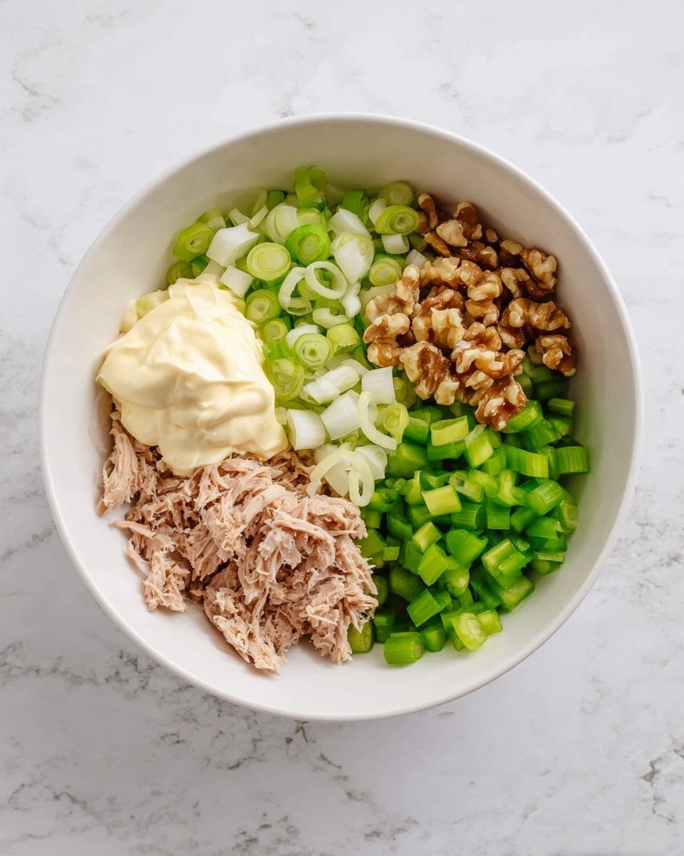 A white bowl sits on a white marbled surface, filled with different ingredients arranged in sections rather than mixed. At the bottom, there are pieces of shredded tuna with a light brown color and flaky texture. To the left, there is a creamy off-white mayonnaise piled in a dollop. Above the mayonnaise, there are thinly sliced green onions with light green and white circular rings. On the top right, there are roughly chopped walnut pieces in a light brown color with a rough texture. Below the walnuts, there is a pile of bright green diced celery with a crisp texture. The ingredients create a clear contrast in colors and textures, arranged in a neat, open layout. Photo taken with an iphone --ar 4:5 --v 7