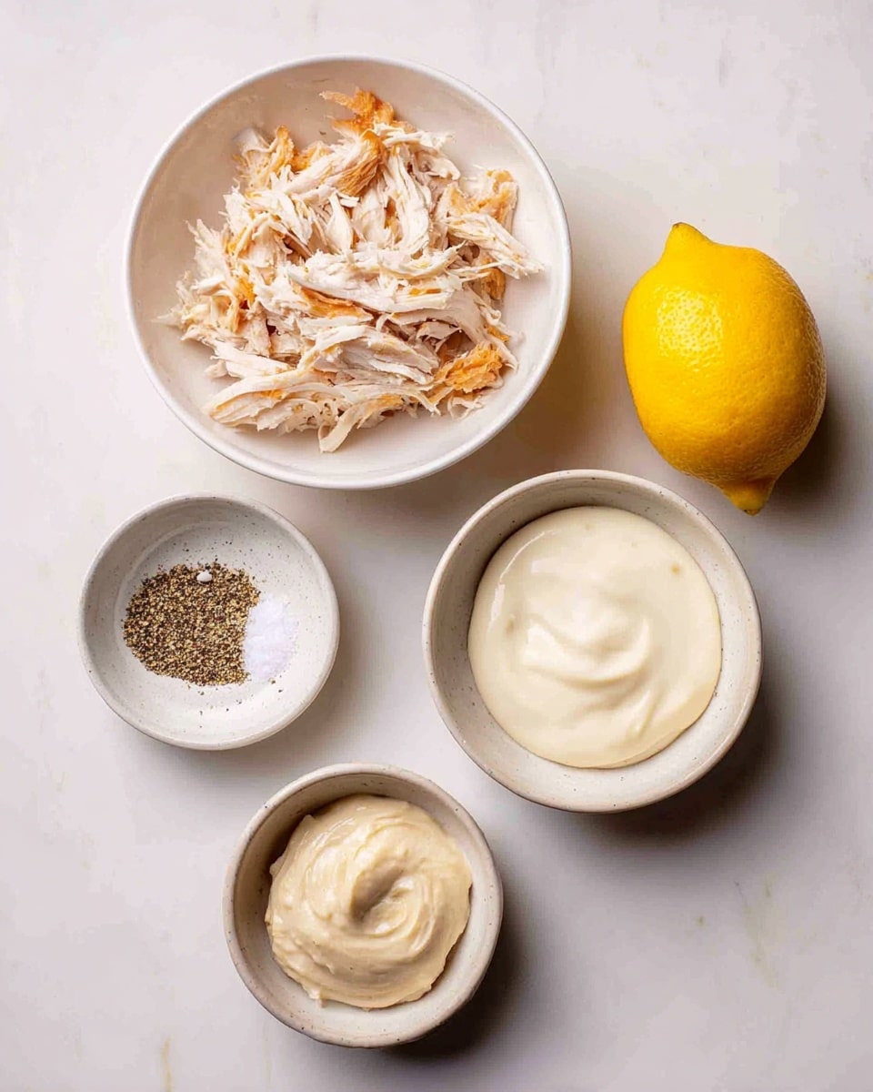 This image shows five food items on a white marbled surface. In the top left, there is a white bowl filled with shredded cooked chicken, showing light brown and white pieces mixed together. To the right of the chicken, there is a whole bright yellow lemon. Below the lemon, a small white bowl holds coarse salt mixed with black pepper. At the bottom left, there is a white bowl filled with smooth, creamy white sauce. Next to it on the right, a small light gray bowl contains a thick, pale, beige-colored paste. The layout is clean and simple, with the bowls and lemon spaced evenly. Photo taken with an iphone --ar 4:5 --v 7