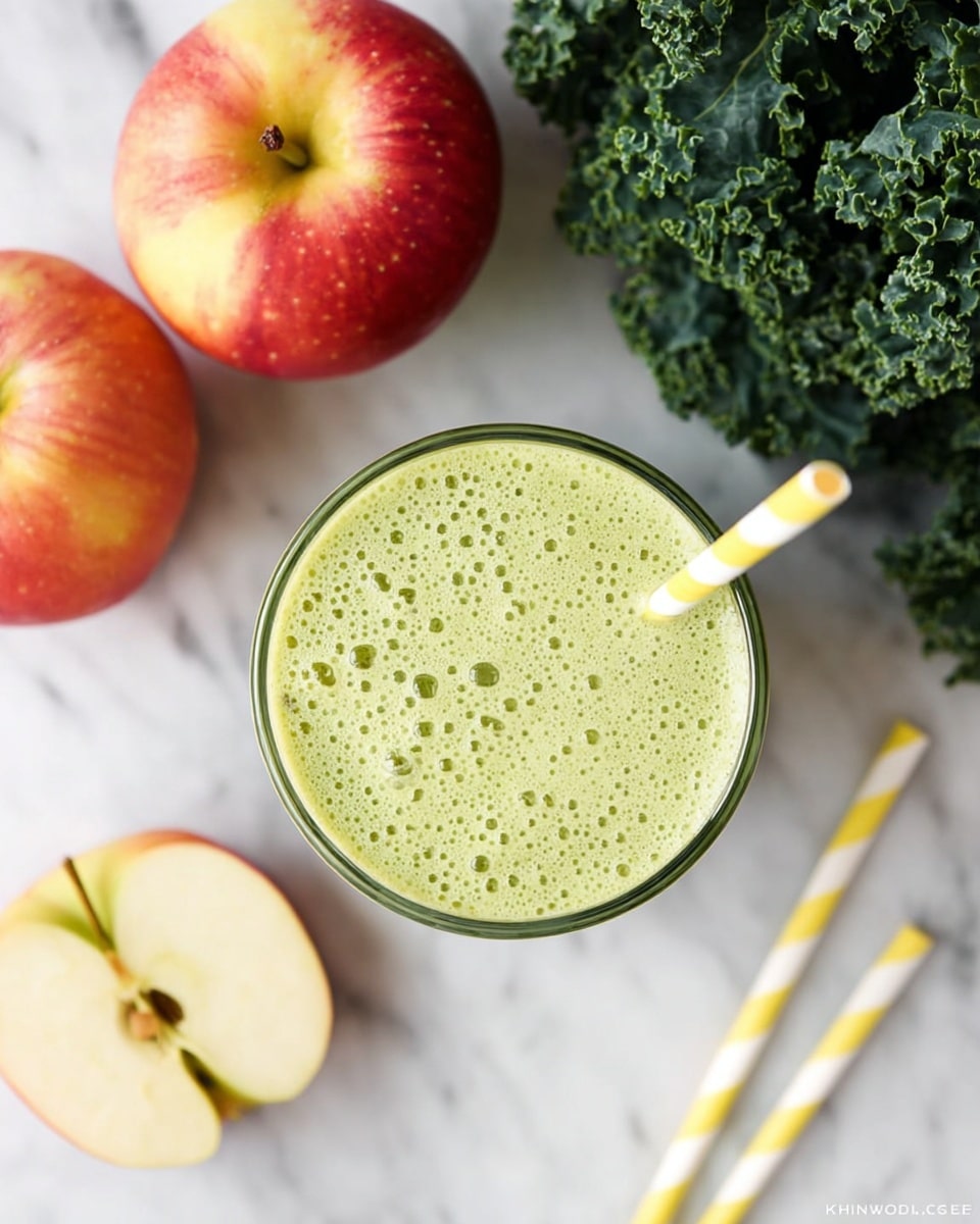 A top view of a clear glass filled with a light green smoothie showing a frothy texture with small bubbles on top. A yellow and white striped straw is placed inside the smoothie near the right edge of the glass. To the left of the glass, three apples are arranged, including one whole red and yellow apple, one apple cut in half showing the light cream inside, and one apple slice. On the top right corner, fresh dark green curly kale leaves are placed. The background is a white marbled surface with two more yellow and white striped straws lying near the bottom right. photo taken with an iphone --ar 4:5 --v 7
