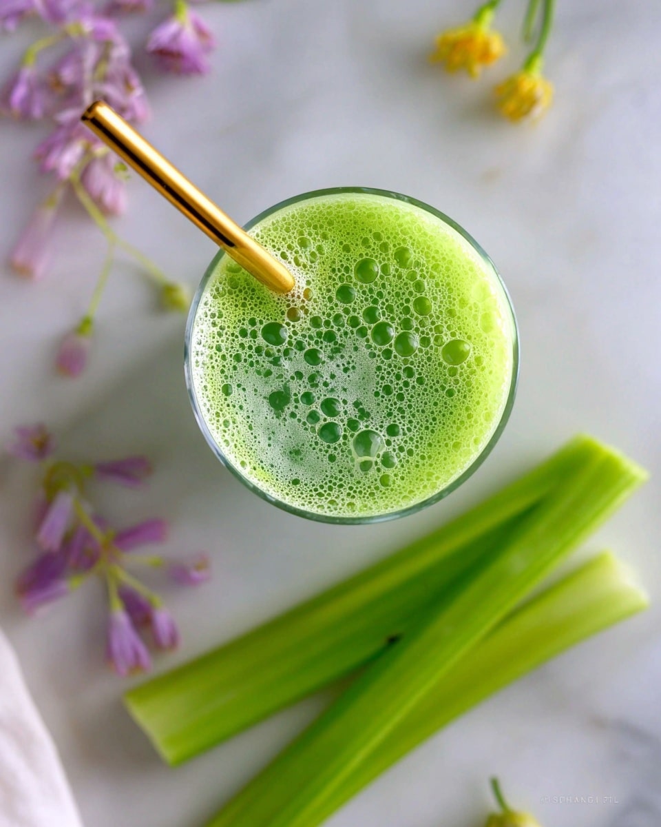 A clear glass filled with bright green juice topped with light green foam and bubbles is shown from above. A shiny golden straw is placed diagonally inside the glass at the top left. In the bottom right corner, there are three long, fresh celery sticks with a glossy texture. The background is a white marbled surface with a soft focus on some small purple and yellow flowers scattered around. Photo taken with an iphone --ar 4:5 --v 7
