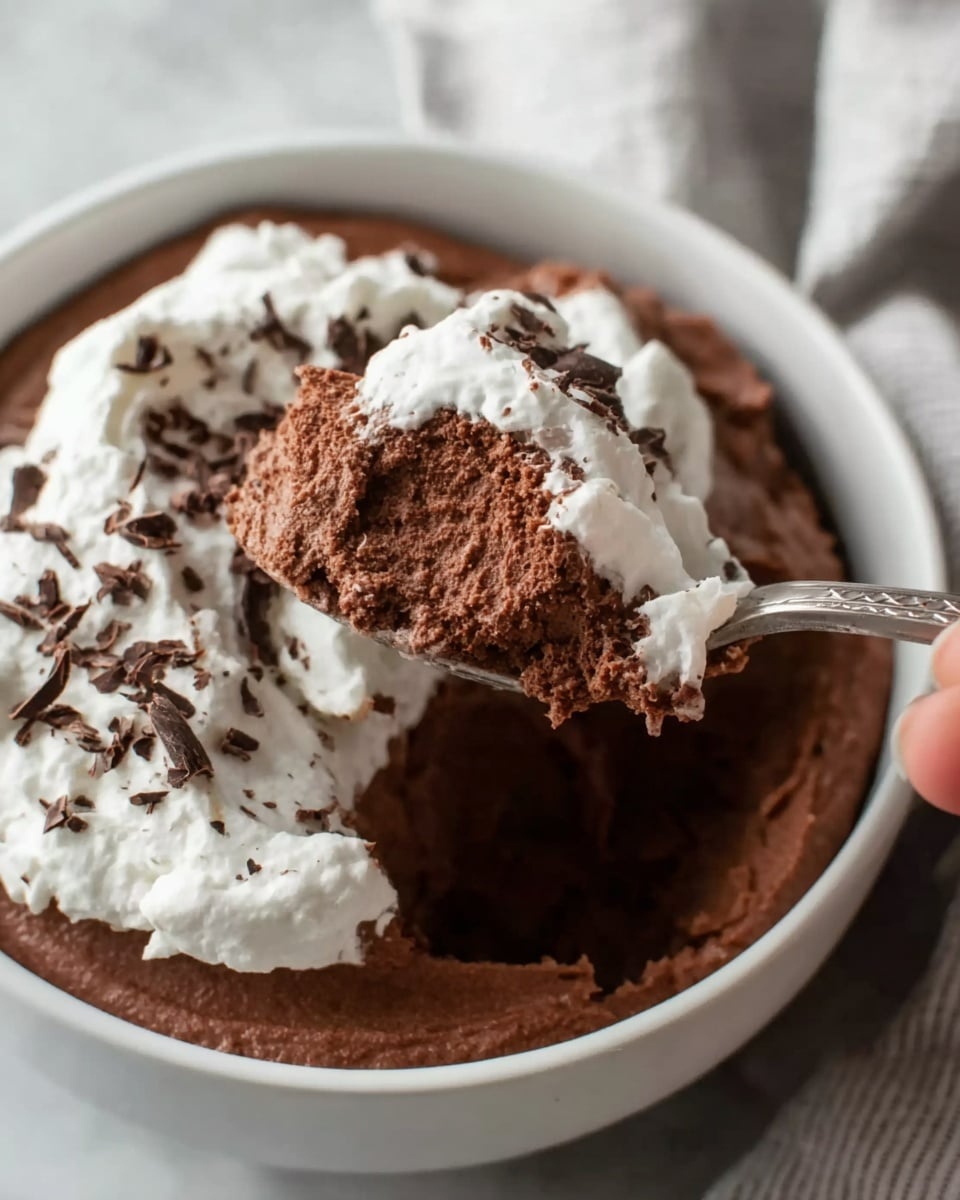 A white bowl filled with two visible layers: a thick, dark brown chocolate mousse on the bottom and a white whipped cream layer on top, both having a soft and fluffy texture. Small dark chocolate shavings sprinkle over the whipped cream, adding a rough texture and extra detail to the top. A spoon scoops a large portion of the chocolate mousse and whipped cream, held by a woman's hand with just the fingers visible. The bowl rests on a white marbled surface with a cloth nearby, creating a cozy and simple setting. photo taken with an iphone --ar 4:5 --v 7