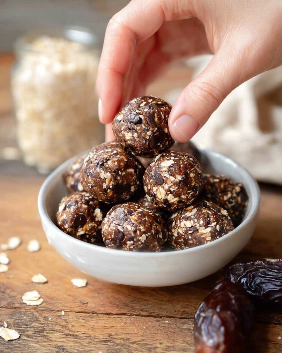 A close-up view of a white bowl filled with around twelve round dark brown energy balls that have visible oats and chocolate chips embedded in them, creating a textured look with light beige and dark spots. A woman's hand is gently lifting one energy ball from the bowl with fingers pinching it near the top center of the image. The bowl sits on a wooden surface with two pieces of shiny dark brown dates near the front right side. The background features a blurred glass container filled with oats and some light-colored objects. The overall setting is casual and warm. Photo taken with an iphone --ar 4:5 --v 7
