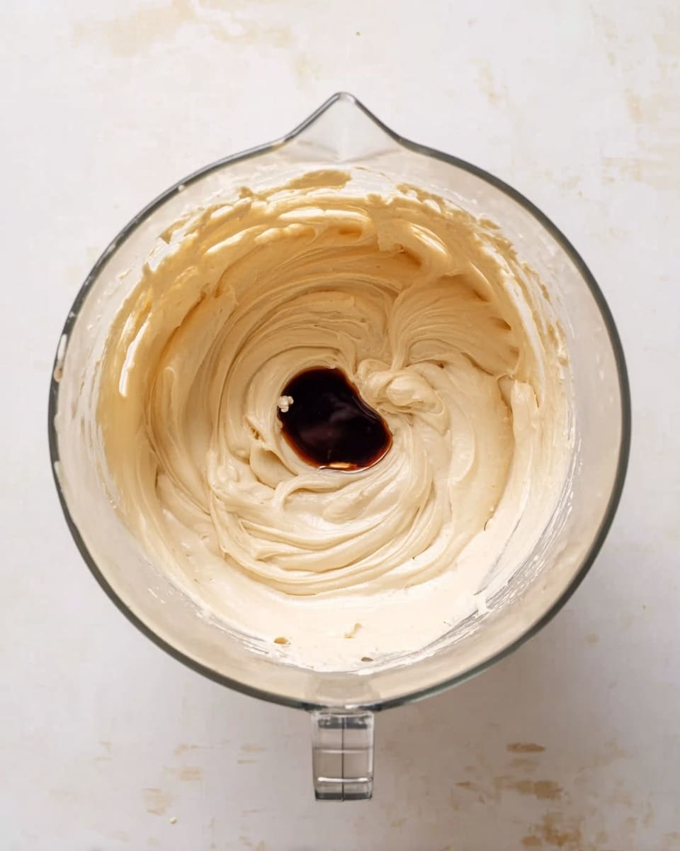 A clear glass mixing bowl sits on a white marbled surface filled with smooth and creamy beige batter that has soft swirls creating a light texture. In the center of the batter, there is a small dollop of dark brown syrup-like liquid sitting atop the creamy layer, contrasting sharply with the pale mixture. The bowl has a handle on one side and a pouring spout on the opposite, and there are traces of batter on the inner sides, showing it is being mixed. The top view highlights the gentle folds and the thick consistency of the batter. photo taken with an iphone --ar 4:5 --v 7
