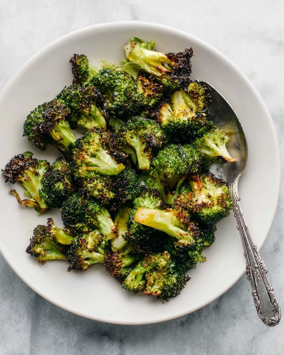 A white plate on a white marbled surface holds a pile of roasted broccoli florets. The broccoli pieces are green with charred, dark brown edges, showing a crispy texture. The florets are irregularly shaped with some stems visible, and a few pieces have a light yellow tint from roasting. A silver spoon with a detailed handle rests inside the plate on the right side, slightly buried under some broccoli. The lighting highlights the colorful contrast and textures of the broccoli. photo taken with an iphone --ar 4:5 --v 7