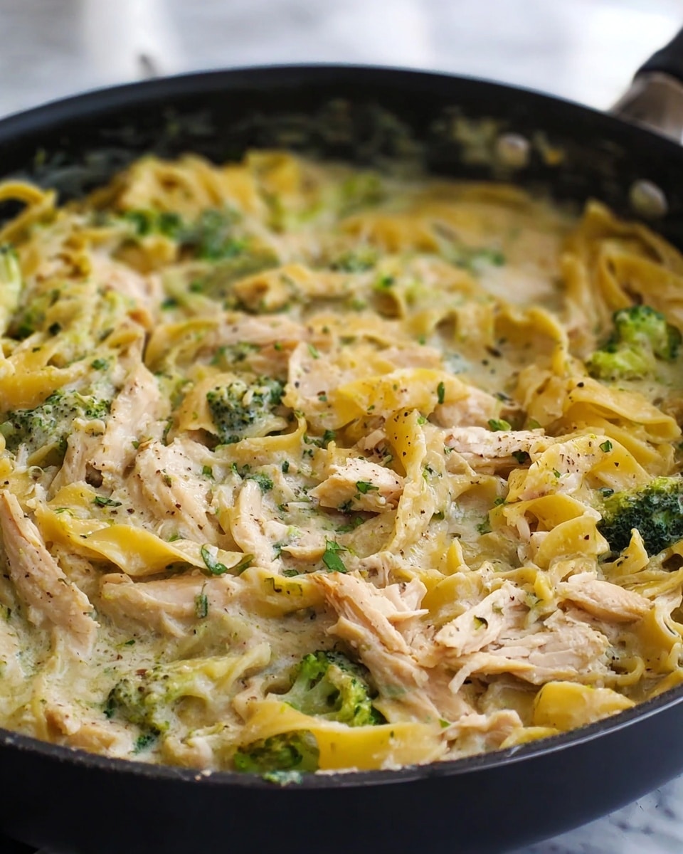The dish shows a creamy pasta with visible layers of yellow twisted noodles mixed with soft, shredded light brown chicken pieces and small bright green broccoli florets evenly spread throughout the creamy white sauce. The creamy sauce has a smooth texture and some black pepper specks are visible on the ingredients. The food is inside a black pan with a slight shine, sitting on a white marbled surface. The image is close up and focused on the mix of pasta, chicken, broccoli, and sauce. photo taken with an iphone --ar 4:5 --v 7