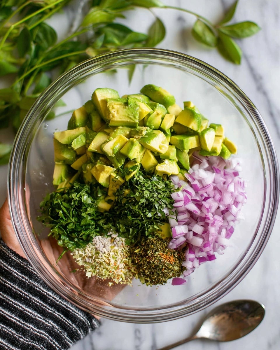 A clear glass bowl on a white marbled surface holds diced green avocado pieces piled at the back, with small clusters of finely chopped purple onion on the right. In front of the avocado and onion, there are small heaps of minced garlic, chopped green herbs, and dried mixed spices arranged neatly side by side. The bowl is held by a woman's hand wearing a black and white striped sleeve. Fresh green leaves peek in from the upper left corner, and a silver spoon lies on the surface below the bowl. photo taken with an iphone --ar 4:5 --v 7