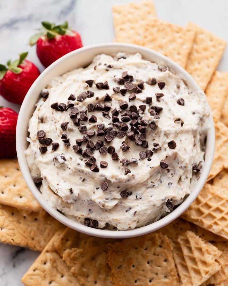 A white bowl filled with a creamy, light-colored mixture that has many small dark chocolate chips spread throughout and sprinkled on top in an even layer, sitting in the center of the image. Around the bowl, there are waffle-like light brown crackers and some light brown square crackers arranged neatly facing outward. On the left side near the crackers, there are two half-cut strawberries with red color and green leaves. The background has a white marbled texture. photo taken with an iphone --ar 4:5 --v 7