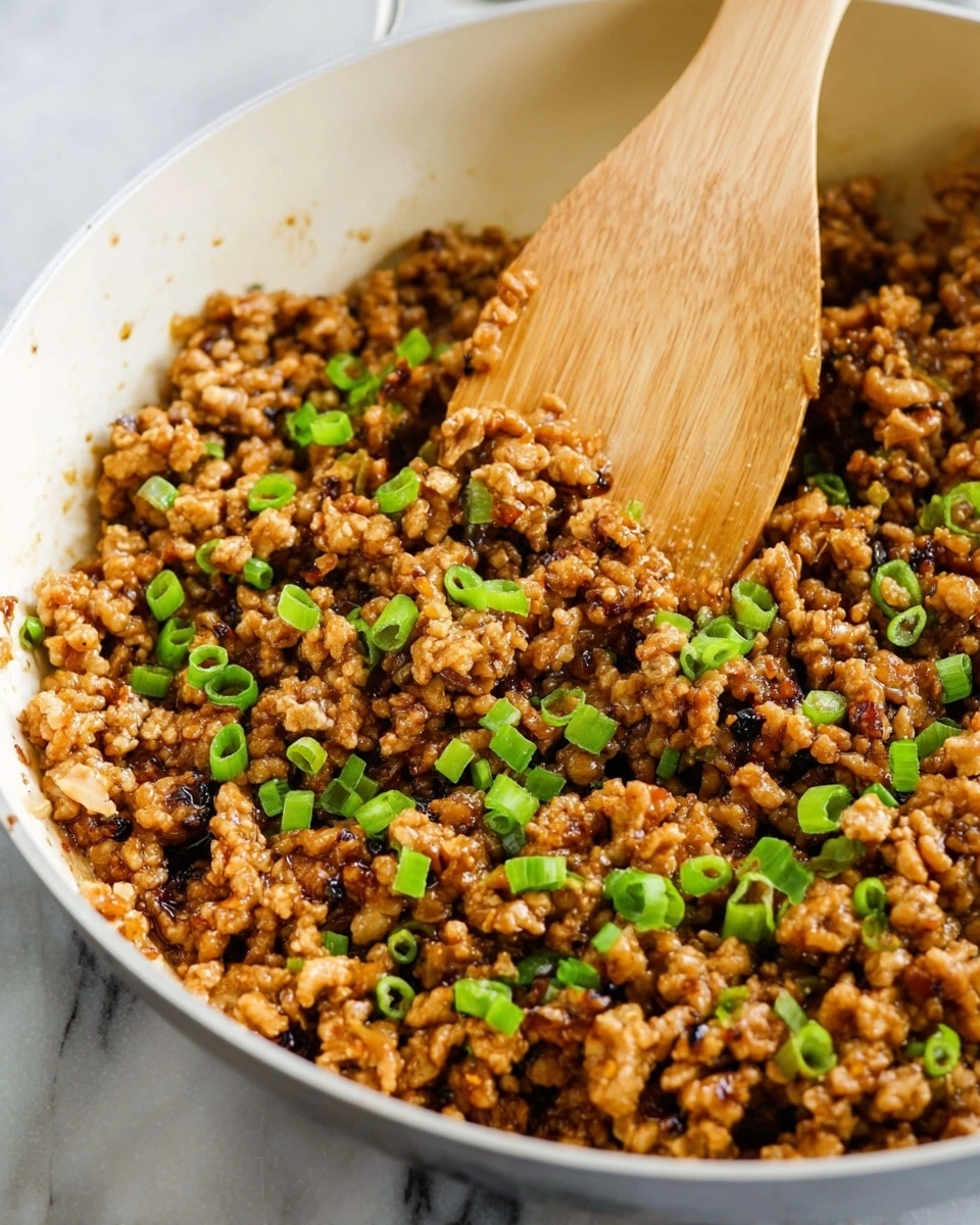 A white pan filled with a cooked minced meat mixture that looks soft and glossy with shades of light brown and darker brown pieces, mixed with small green chopped spring onions sprinkled evenly on top. A wooden spatula is stirring the mixture, showing the textured and slightly shiny surface of the meat and vegetables. The background is a white marbled texture. Photo taken with an iphone --ar 4:5 --v 7