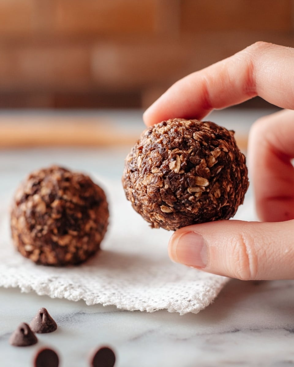 The image shows two round chocolate oat balls with a rough texture, made of oats and small chocolate pieces. One ball is close to the camera, held gently between thumb and forefinger of a woman's hand on the right side, showing the textured, dense mix with visible oats and chocolate bits. The second ball is placed on a folded white paper towel in the background on a white marbled surface. Small chocolate chips are scattered around the paper towel, and the background includes a blurred brown surface and brick wall. photo taken with an iphone --ar 4:5 --v 7