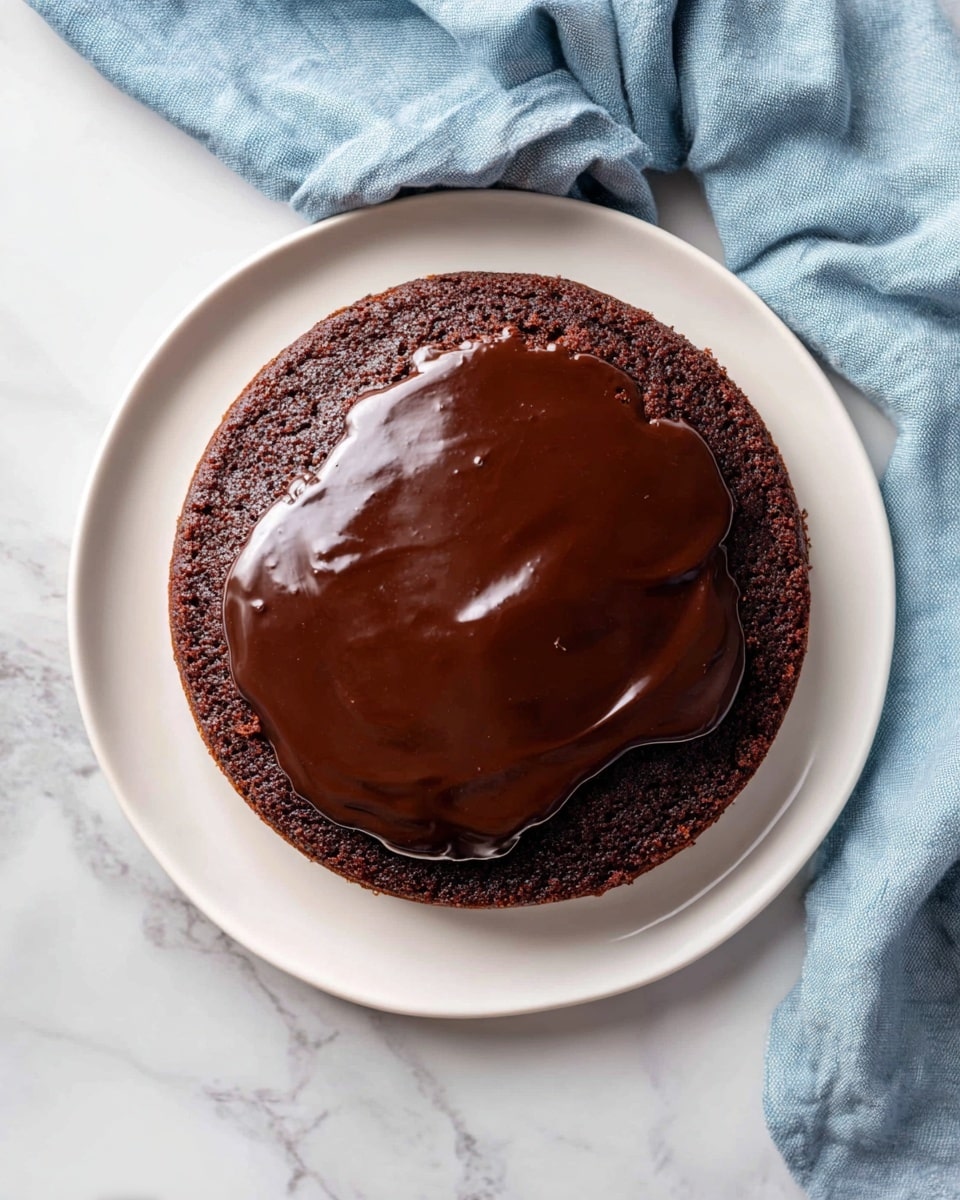 A single round chocolate cake layer sits flat on a white plate. On top of the cake is a thick, shiny layer of dark chocolate glaze that spreads unevenly, mostly covering the center but leaving some cake edges visible. The cake has a soft, slightly crumbly texture and deep brown color, while the glaze is smooth and glossy with a rich dark brown hue. The plate rests on a white marbled surface with a soft blue cloth draped casually to the left side. photo taken with an iphone --ar 4:5 --v 7