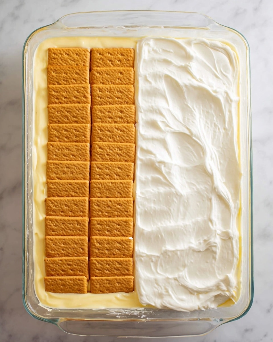 The image shows a dessert in a clear glass baking dish placed on a white marbled surface. The dessert has two main layers. In the left part, the bottom layer is creamy yellow, smooth, and thick, topped with a neat layer of rectangular golden brown crackers arranged in three rows and four columns. In the right part, there is a thick, smooth white cream layer spread evenly all over the top, looking soft and swirled gently with a spatula. The glass dish is rectangular with rounded edges, and the dessert fills it almost to the top. Photo taken with an iphone --ar 4:5 --v 7