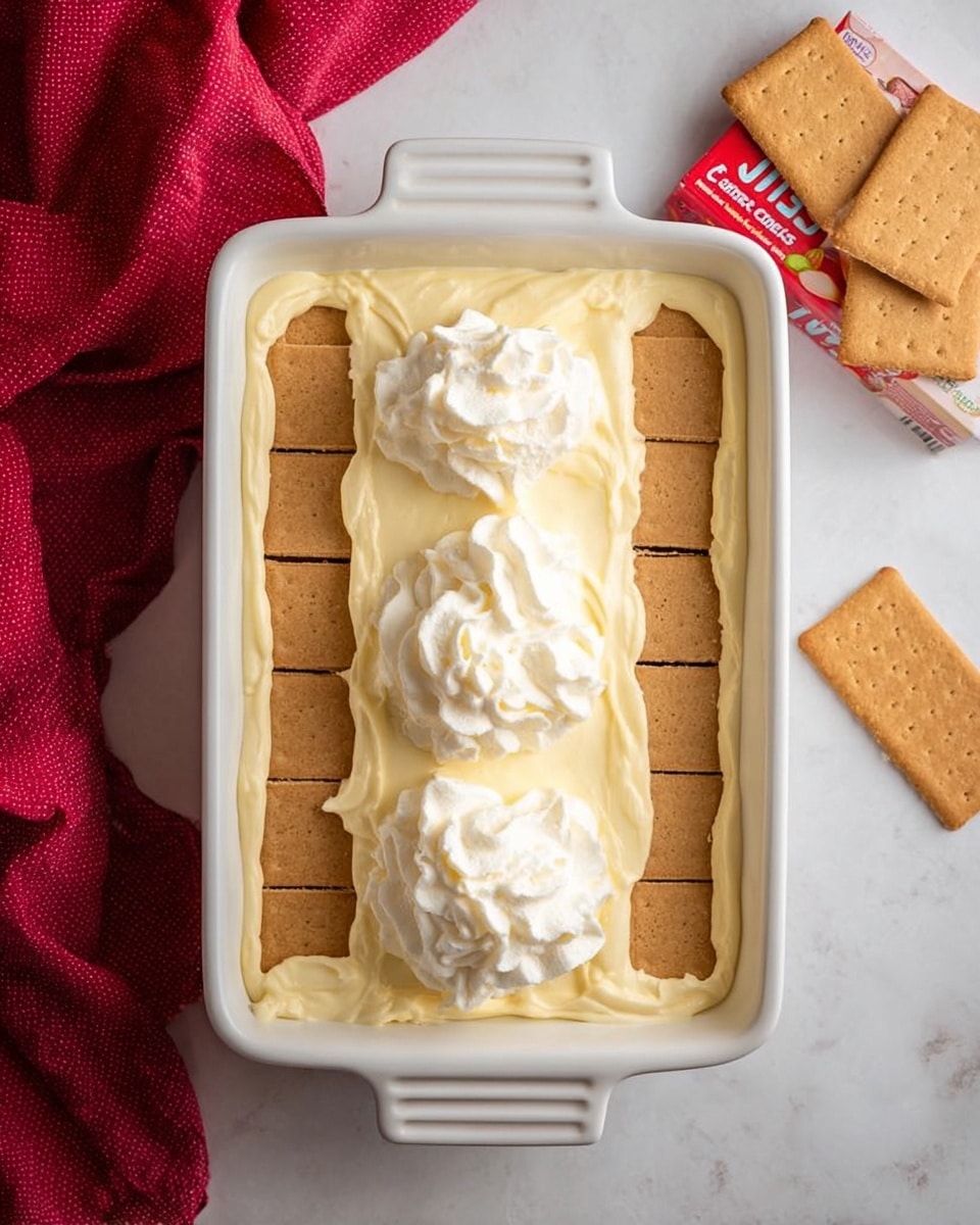 The image shows a white rectangular dish filled with a grid of light brown graham cracker squares arranged evenly in one layer at the bottom. On top of the crackers, there is a thick, creamy pale yellow layer of whipped pudding dolloped in three large sections down the center. To the top right of the dish, there are three more graham crackers placed in a neat stack on a white marbled surface. A crumpled red cloth lies to the left of the dish, and two pudding mix boxes sit near the bottom right corner of the frame. Photo taken with an iphone --ar 4:5 --v 7