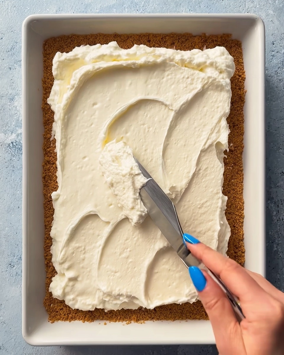 The image shows a close-up of a square dessert base with a golden brown crumb crust layer, evenly spread in a white rectangular dish. On top of the crust, there is a thick, creamy white layer being smoothed out with a metal spatula held by a woman's hand with blue nail polish. The creamy layer looks soft with light air bubbles, and its texture is smooth but slightly uneven from the spreading motion. Photo taken with an iphone --ar 4:5 --v 7