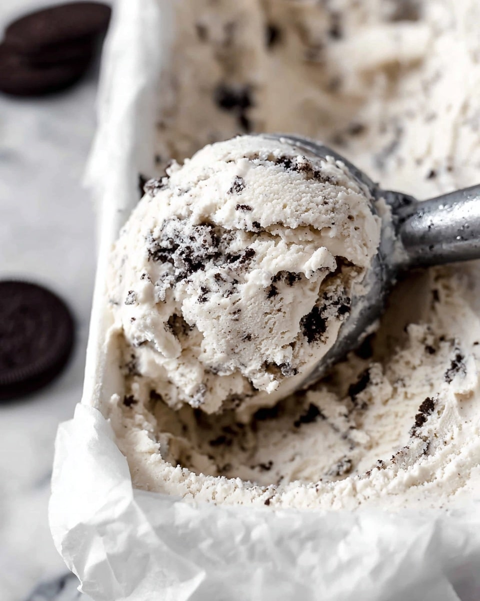 A close-up view of a scoop of cookies and cream ice cream with a creamy white base filled with small dark chocolate cookie bits, held by a silver ice cream scoop. The ice cream sits in a white container lined with white parchment paper, showing soft, textured ice cream with visible cookie crumbs throughout. The background features a white marbled surface with a few dark chocolate cookies slightly blurred in the back. photo taken with an iphone --ar 4:5 --v 7